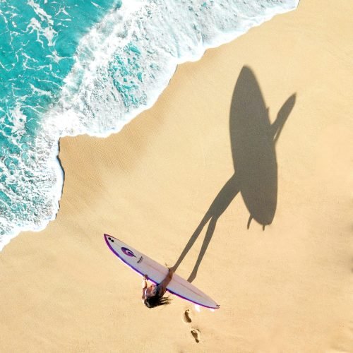Aerial shot of a woman with a surfboard casting a shadow on a sandy beach in Haleiwa, Hawaii.